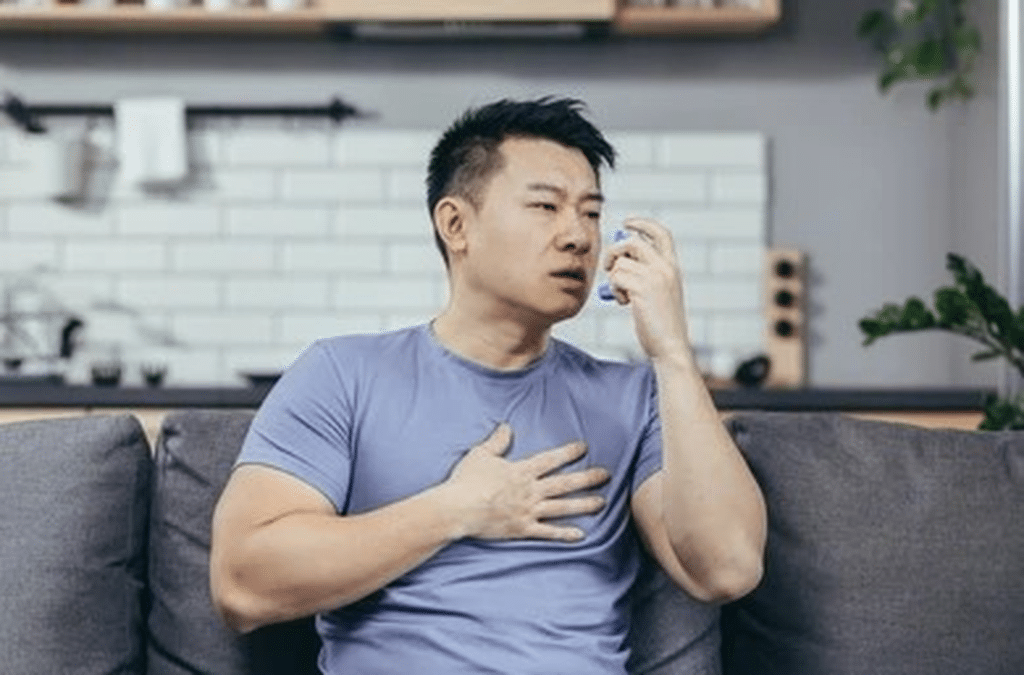 Man sitting on a couch and using an inhaler to treat his asthma symptoms.